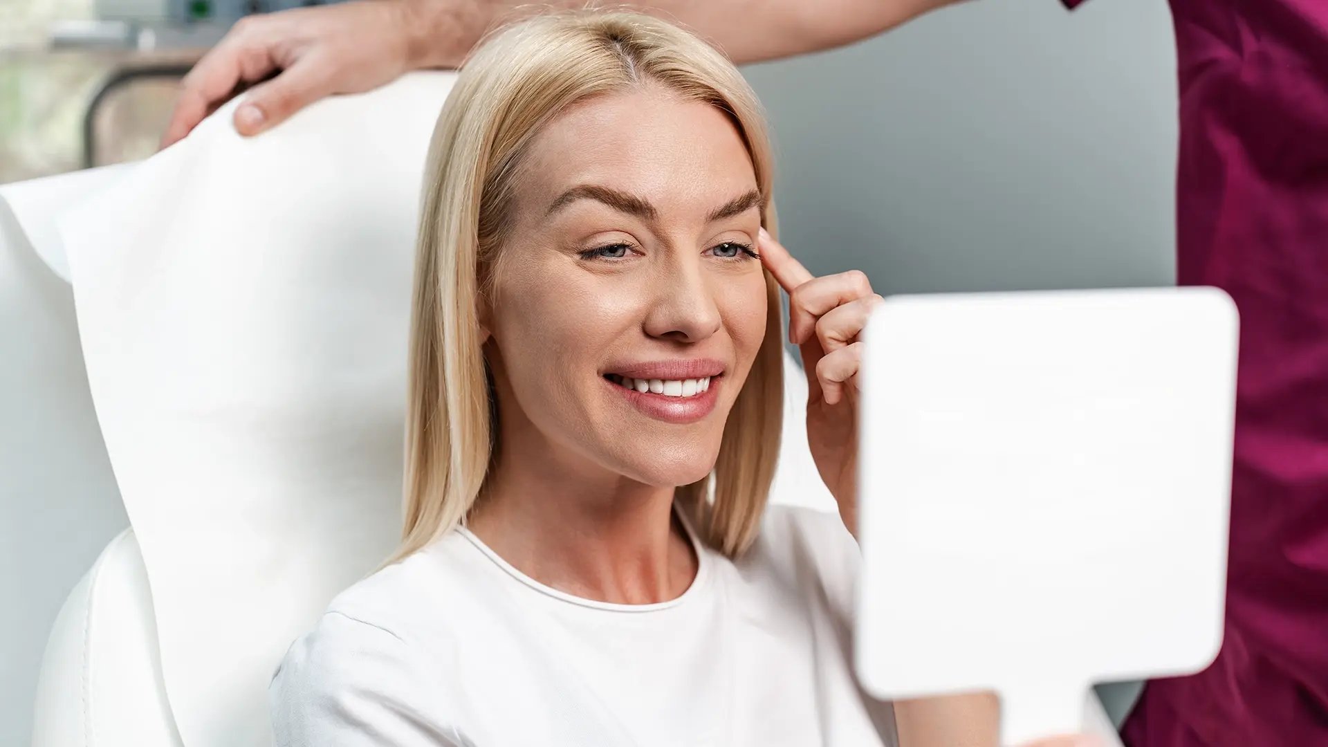 Blond woman smiles at her reflection in a mirror after getting a Jeuveau treatment at Liquivida Wellness Center